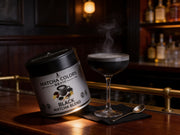 Lata de Black Matcha Blend junto a una copa coupe con bebida negra y humo en un bar speakeasy de luz ámbar - campaña IA