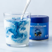 Cuchara de polvo azul de Blue Matcha Blend creando remolinos oceánicos en un vaso de leche, lata detrás - campaña IA