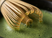 Macro de las puas rizadas del chasen de bambu de Matcha Colors emergiendo de la espuma de matcha - campaña IA