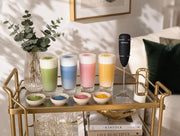 Espumador electrico de Matcha Colors en un carrito bar dorado junto a lattes de colores verde azul rosa y amarillo - campaña IA