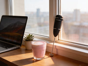 Espumador de leche electrico de Matcha Colors junto a un latte rosa en un escritorio de home office con luz de manana - campaña IA