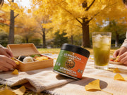Lata de Genmaicha Matcha Premium en un picnic de otoño con bento y té helado bajo los ginkgos - campaña IA
