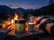 Lata de Hojicha Matcha Premium junto a una fogata de montaña al anochecer, con taza esmaltada de hojicha latte y manta de lana - campaña IA