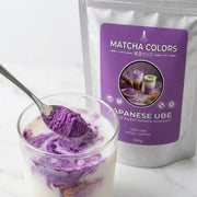 Cuchara de polvo de Japanese Ube creando remolinos morados en un vaso de leche, doypack de Matcha Colors detrás - campaña IA