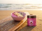 Smoothie bowl rosa de pitaya sobre una tabla de surf en la arena al amanecer, con la lata de Pink Pitaya Powder al lado - campaña IA