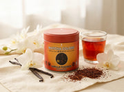 Lata de Vanilla Rooibos Powder de Matcha Colors entre orquídeas blancas, vainas de vainilla y té rooibos con luz crema suave - campaña IA