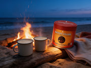 Lata de Vanilla Rooibos Powder de Matcha Colors junto a tazas esmaltadas ante una hoguera en la playa al anochecer - campaña IA