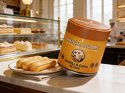 Lata de Vanilla Chai Blend de Matcha Colors sobre el mostrador de mármol de una pastelería francesa junto a éclairs de vainilla - campaña IA